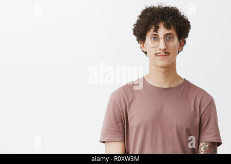 Taille - geschossen von freundlich - suche Ruhe und unbeschwerten jungen Mann mit Schnurrbart und lockige Frisur, Brille und braunes t-shirt lächelnde niedliche Ständigen beiläufig über grauer Hintergrund Stockfoto