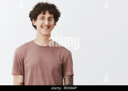 Studio shot von charmanten motiviert gut aussehende lockigen dunkelhaarigen Mann mit Schnurrbart und tätowiert arm in Gläsern und trendy t-shirt freudig lächelnd und freundlich Gefühl erreicht bis zu Träume Stockfoto