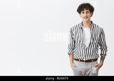 Taille-up Shot der stilvolle, moderne junge Hipster Kerl in gestreiften Hemd und graue Hosen, Hände in den Taschen mit Schnurrbart dunkle lockige Haare und Tattoos Dressing up für Datum über grauer Hintergrund Stockfoto