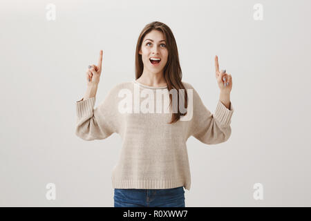 Mädchen aufgeregt wird ihr Name im Himmel geschrieben sehen. Portrait der expressiven schöne Frau mit langem Haar suchen und zeigt mit Index Finger, lächelnd, keuchend von positiven Überraschung über graue Wand Stockfoto