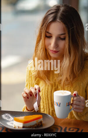 Hübsche junge Frau genießen eine Tasse Kaffee und ein Stück leckeren Kuchen beim Stillstehen, die Sonne scheint durch die Fenster im Cafe. Stockfoto