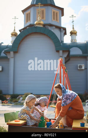 Russisch-orthodoxe Frau in einem Schal spielen mit ihren zwei Töchtern auf dem Spielplatz auf dem Hintergrund der russisch-orthodoxen Kirche aus Holz. Stockfoto
