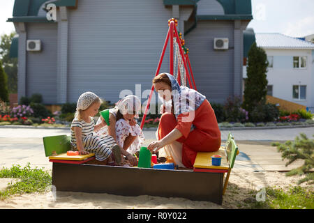 Russisch-orthodoxe Frau in einem Schal spielen mit ihren zwei Töchtern auf dem Spielplatz auf dem Hintergrund der russisch-orthodoxen Kirche aus Holz. Stockfoto