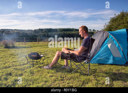Kaukasische Mann erholsamen Abend genießen Sie ein kühles Bier durch seine Feuerstelle ausserhalb seiner blauen Zelt in einem Feld genießt die Aussicht Stockfoto