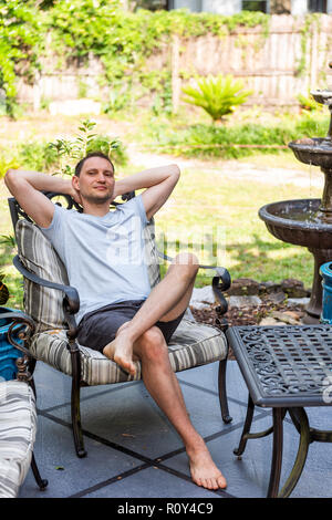 Junge glücklicher Mann sitzen auf der Terrasse Lounge Chair in outdoor Frühling Blumen Garten im Hinterhof des Hauses zen mit Springbrunnen, Tisch, Pflanzen Stockfoto