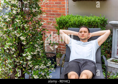Junge glücklicher Mann sitzen auf der Terrasse Lounge Chair in outdoor Frühling Blumen Garten im Hinterhof des Hauses zen, Pflanzen Stockfoto