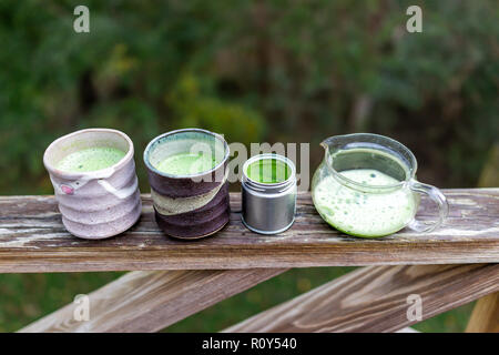 Teekanne aus Glas auf hölzernen Geländer auf Hinterhof deck Garten, zwei Tassen mit Japanischen pulsierenden grünen Tee matcha Farbe gefüllt, Schaum in Morgen Stockfoto