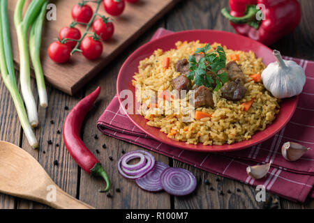 Rote Platte mit Pilav auf braunem Holz- Tabelle. Auf dem Tisch, rote Paprika, grüne Zwiebeln, Knoblauch, Tomaten, rote Serviette und Löffel. Stockfoto