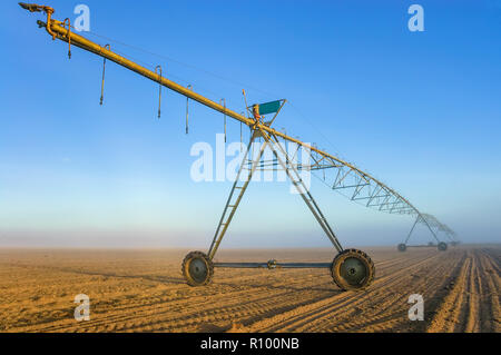 Die aufgehende Sonne langsam verbrennt der Nebel aus den Furchen des Feldes nach und nach Aufdecken des Regnerauslegers bereit die Bewässerung der Pflanzen Saatgut zu beginnen. Stockfoto