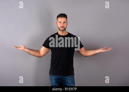 Ein hübscher junger Mann in einem schwarzen T-Shirt, die etwas mit seinen Handflächen vor einem grauen Hintergrund im Studio. Stockfoto