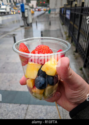 Pov Nahaufnahme von Hand hält transparenten Kunststoff Tasse geschnittene Früchte, Beeren, Erdbeere, Ananas, Mango. Gesunde Street Food Snack Stockfoto