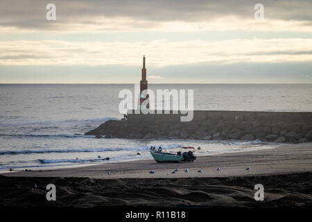 Ein Fischer und einem kleinen Boot sitzen in der Nähe ein leuchtfeuer an einem Strand. Blick auf das Meer, bewölkten Tag. Stockfoto