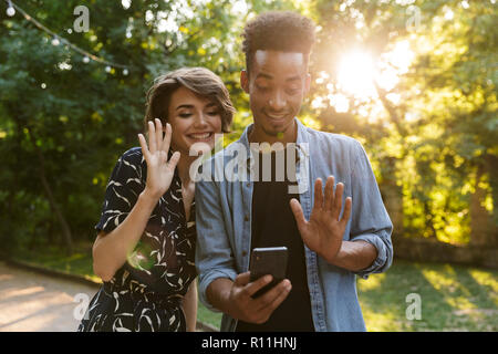 Happy multiethnischen Paar im Park, winkende Hände zu Mobiltelefon Stockfoto