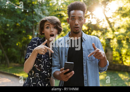 Überrascht multiethnischen Paar im Park, der auf Handy Stockfoto