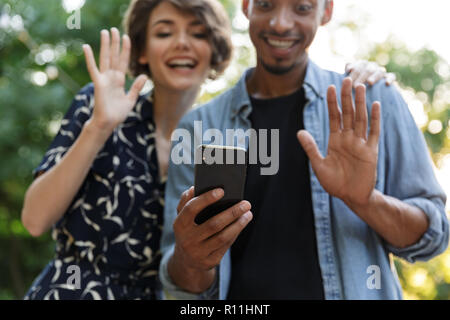 Zugeschnittenes Bild der glücklichen multiethnischen Paar im Park, winkende Hände zu Mobiltelefon Stockfoto