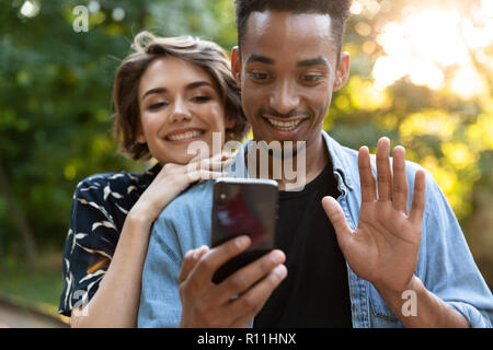 Lächelnd multiethnischen Paar im Park, winkende Hände zu Mobiltelefon Stockfoto