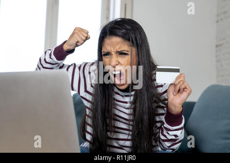Attraktive junge Latin Frau verzweifelt und wütend mit Kreditkarte und Laptop Standortwahl auf dem Sofa zu Hause in der Kreditkarte Probleme Schulden online Zahlung online Stockfoto