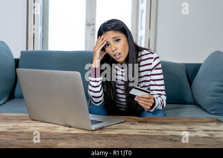 Attraktive junge Latin Frau verzweifelt und wütend mit Kreditkarte und Laptop Standortwahl auf dem Sofa zu Hause in der Kreditkarte Probleme Schulden online Zahlung online Stockfoto