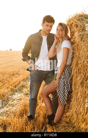 Volle Länge Foto, junges Paar, Mann und Frau zu Fuß auf dem Feld und in der Nähe von großen Heuhaufen Stockfoto