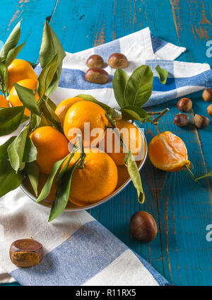 Flach auf reife Orangen Mandarinen Obst mit grünen Blättern, in eine Schüssel, und Kastanien, über rustikale blau Holz- Hintergrund, Vertikal, kopieren. Stockfoto