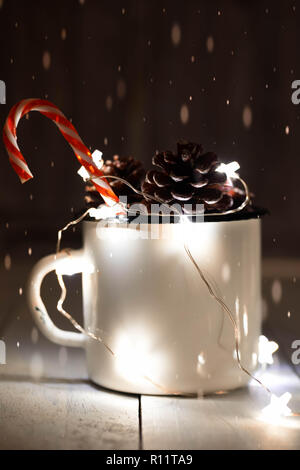 Tannenzapfen in Zinn Glas mit funkelnden sternförmige Garland und einzelne Zuckerstange stick. Weihnachtsschmuck. Dark Photo. Fallenden Schnee Effekt. Stockfoto