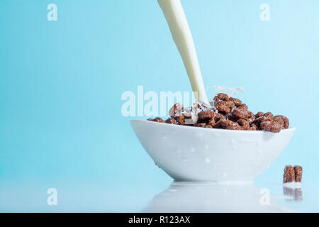 Milch stream jet Gießen in eine Schale mit Schokolade Flocken in der Form von Ringen, Spritzer von Milch auf einem hellblauen himmlischen Hintergrund Stockfoto