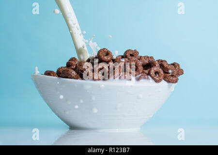 Milch stream jet Gießen in eine Schale mit Schokolade Flocken in der Form von Ringen, Spritzer von Milch auf einem hellblauen himmlischen Hintergrund Stockfoto