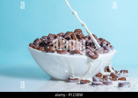 Milch stream jet Gießen in eine Schale mit Schokolade Flocken in der Form von Ringen, Spritzer von Milch auf einem hellblauen himmlischen Hintergrund Stockfoto