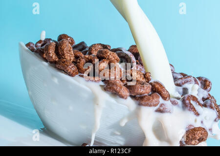 Milch stream Gießen in eine Schale mit Schokolade Flocken in der Form von Ringen, Spritzer von Milch auf einem hellblauen Hintergrund Stockfoto