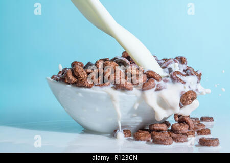 Milch stream jet Gießen in eine Schale mit Schokolade Flocken in der Form von Ringen, Spritzer von Milch auf einem hellblauen himmlischen Hintergrund Stockfoto