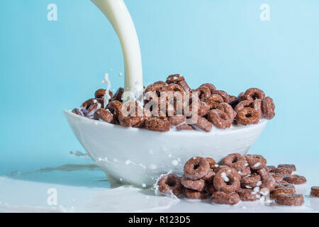 Milch stream jet Gießen in eine Schale mit Schokolade Flocken in der Form von Ringen, Spritzer von Milch auf einem hellblauen himmlischen Hintergrund Stockfoto