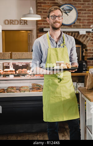 Porträt einer Mitte nach Kellner in einem Coffee Shop. er in die Kamera schaut und lächelnd, während Sie ein Tablett mit Kaffee und Scone. Stockfoto