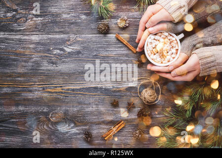 Frau und Mann die Hände halten Sie die Tasse heißen Kakao mit Marshmallows auf Holzuntergrund mit pine Äste, Tannenzapfen, Zimtstangen und Anis Sterne mit Lichtern geschmückt. Winter heiß trinken. Stockfoto