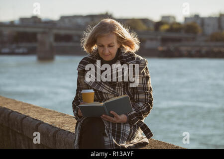 Ältere Frau genießt Buch lesen und Kaffee trinken, während Sie durch den Fluss sitzen. Stockfoto