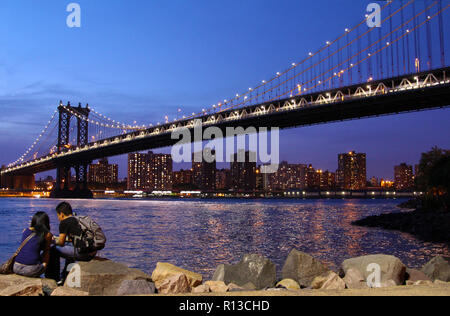 Jungen asiatischen Paar sitzt auf Felsen vor Manhattan Bridge ab, die im Rahmen der Manhattan Bridge Überführung (Dumbo), New York, NY gesehen Stockfoto