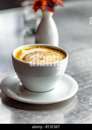 Casual Coffee Shop. Latte Art schäumenden Latte. Wasser Kanne in das klare Glas. orange Blumen in eine weiße, ovale weiße Vase. Stockfoto