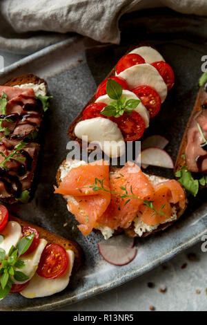 Sandwich mit Roastbeef und Rucola, Lachs und Kreuzkümmel, Mozzarella und Tomaten auf braunem Brot am grauen Platte Stockfoto