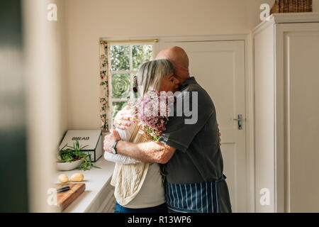 Älterer Mann umarmt seine Frau steht in der Küche. Ältere Menschen, seine Liebe zu seiner Frau, die Blumen und eine herzliche Umarmung. Stockfoto