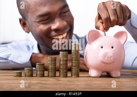 Happy afrikanischer Mann einfügen Münze in Sparschwein in der Nähe von Stapel von Münzen Stockfoto