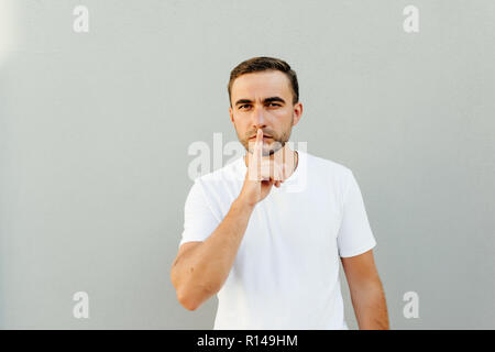 Shhh Geste. Junger Mann auf grauem Hintergrund in Weiß gekleidet casual t-shirt Drücken mit dem Finger auf die Lippen, als ob sie Stille zu halten. Stockfoto