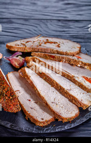 Schweinebraten, beschichtet mit roten Gewürze, in Scheiben schneiden, Knoblauchzehen, serviert auf einem schwarzen Stein Board auf einem schwarzen Holztisch, vertikal anzeigen, kopieren Raum Stockfoto