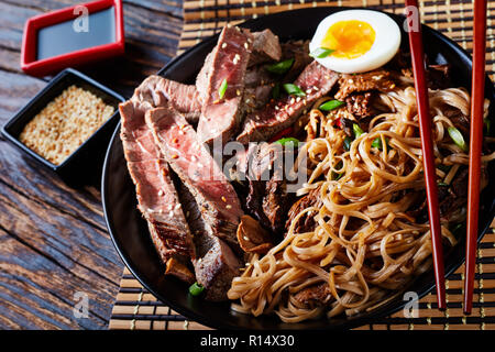 Eine Schüssel mit Soba mit in Scheiben Roastbeef, Shiitake-pilze, die Hälfte der gekochten Ei und Gemüse mit Stäbchen auf einem Bambus Tisch mat. Soja Sauce auf einem Rus Stockfoto