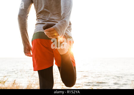 Rückansicht Foto von gutaussehenden jungen Sport Mann stand am Strand machen Dehnübungen. Stockfoto