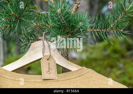 Organische Kleidung. Natürliche farbige T-Shirt hängen auf hölzernen Kleiderbügel im Kiefer. Eco Textile Tag. Grüne wald, natur im Hintergrund. Stockfoto