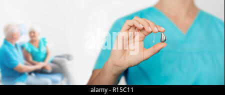 Arzt zeigt ein Hörgerät der letzten Generation mit unfokussierten älteren Patienten im Hintergrund Stockfoto