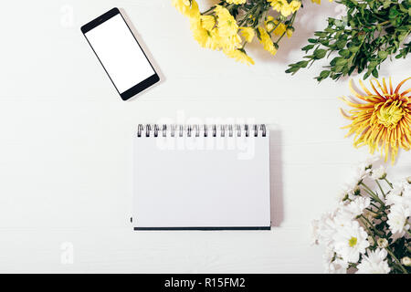 Ansicht von oben Vorlage öffnen leere Notepad, Handy mit leeren weißen Bildschirm unter flach Rahmen von Blumen und grüne Zweige. Stockfoto