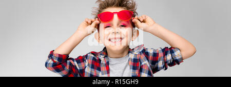 Ein schöner Junge in einem Plaid Shirt, grau Shirt und Jeans steht auf einem grauen Hintergrund. Ein Junge in rot Sonnenbrille. Der Junge hält seine Hand Gläser Stockfoto