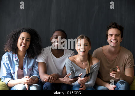 Gerne diverse Studenten multirassischen Freunden zusammen sitzen Indoor. Attraktive lächelnde Mädchen stattliche Kerle holding Mobiltelefone lachend auf c Stockfoto