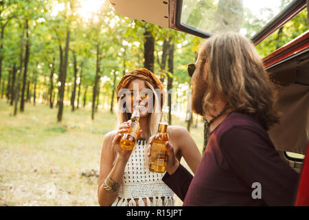 Foto der Schönen hippie Paar Mann und Frau lächelnd und trinken Bier im Wald in der Nähe von Retro minivan Stockfoto