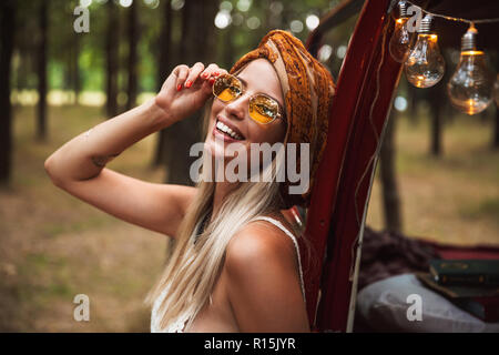 Foto der hübschen Hippie Frau mit stilvollen Accessoires, lächelnd, während in Forest Camp ruhen Stockfoto